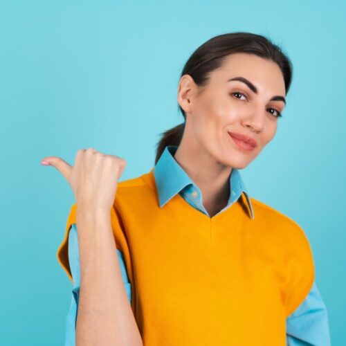 Young woman in a shirt and an orange vest on a turquoise background smiling cheerfully, pointing her finger to the left to an empty space